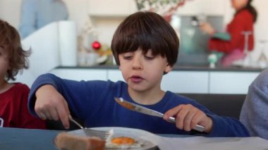 Evde mutfak masasında ailesiyle kahvaltı ederken elinde çatal ve bıçakla sahanda yumurta çiğneyen küçük çocuk.
