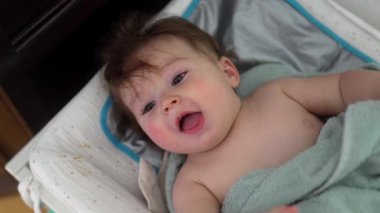 Bebek havluya sarılı bir şekilde masanın üzerinde yatıyor, geniş ağızlı, neşe dolu bir kumaşı ısırıyor, keşif, rahatlık ve çocukluk ifadesi..