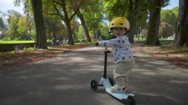 Yeşil kask takmış, yeşil park yolunda scooter süren bir çocuk. Arka manzara kararlılığını gösteriyor. Ağaçlarla kaplı, yaz öğleden sonra.