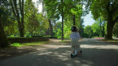 Miğferli küçük bir çocuk gölgeli ağaç çizgili yolda scooter sürüyor, enerjiyle ileri itiyor ve açık havada doğal çevreyi keşfederken odaklanıyor.