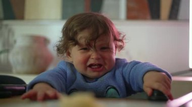 Mavi kıyafetli bir çocuk, kızgın bir ifadeyle yüksek sandalyede oturuyor ve evde aile yemeği sırasında dışarı çıkmak istiyor..