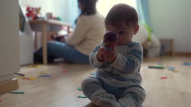 Bebek yerde oturup oyuncağı ağzına koyarken, oyun zamanı boyunca aileyle birlikte günlük ev ortamında merakını ve keşiflerini dile getiriyor..