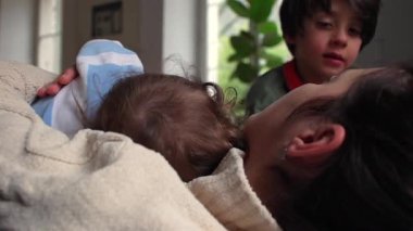 Anne, bebeği göğsünde dinlenirken rahatlatıyor. Büyük kardeş, yakınına eğiliyor. Şefkat, sıcaklık ve duygusal bağ gösteriyor.