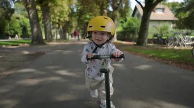 Yeni yürümeye başlayan çocuk park yolunda skuter sürerken sarı kask takıyor, odaklanmış ifade, ağaçlar ve yeşilliklerle çevrili, açık hava keşfi ve bağımsızlığın tadını çıkarıyor.