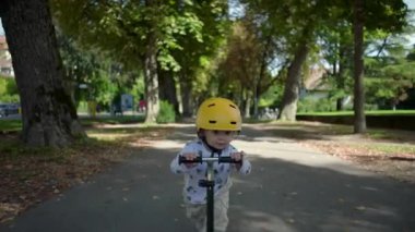 Park yolunda scooter süren küçük bir çocuk sarı kask takıyor, odaklanmış ve kararlı, ağaçlarla çerçevelenmiş, yerde yapraklar, huzurlu bir açık hava.