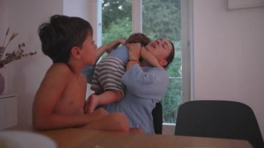 Anne, yeni yürümeye başlayan çocuğu emzirirken yemek masasında büyük kardeşiyle konuşuyor. Ebeveynlik sorunlarını, eğitimi ve sabrı dengelerken hafif bir rahatsızlık ifadesi gösteriyor.