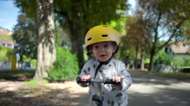 Parkta motosikletle gezen mutlu bir çocuk. Sarı kask takıyor, yakın çekimde gülümsüyor. Güneşli bir günde ağaçlarla ve yeşilliklerle çevrili.