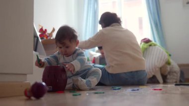 Anne arka planda, merak, oyunculuk ve çocukluk gelişimini gösteren bir anne ile aile yatak odasında otururken yerde bebek keşfi yapan oyuncak fıçısı.