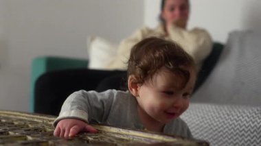 Küçük çocuk masaya yaslanırken gülümsüyor, anne arka planda koltukta oturuyor, günlük ebeveynlik sahnesi nemizi, masumiyetimizi ve erken gelişmemizi gösteriyor..