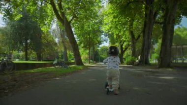 Miğferli bir çocuğun gölgeli yolda scooter sürüşü, küçük bir çocuğun dışarıda azim ve bağımsızlıkla ilerleyişi.