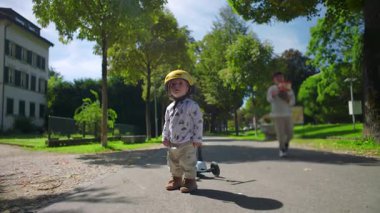 Yeni yürümeye başlayan çocuk dışarıda kask, merak ve masumiyet takarak park yolunda duruyor. Aile sıcaklığıyla arka plandan yaklaşan ebeveyn.