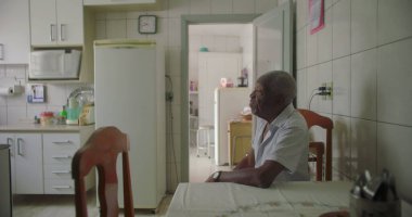 Bir yaşlı siyah adam mutfak masasında oturmuş, kollarını dinlendiriyordu. Etrafı fayanslarla, duvarlarla, buzdolabıyla ve rahat bir ev ortamında ahşap sandalyelerle çevriliydi.