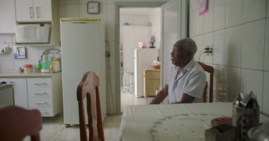 Bir yaşlı siyah adam mutfak masasında oturmuş düşünceli ve düşünceli bir yüz ifadesiyle, döşeli bir iç döşeme, buzdolabı ve bir ev düzeninde ahşap sandalyelerle çevrili.