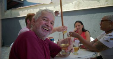 Yaşlı bir adam gülümsüyor ve arka bahçedeki barbekü partisinde bardaktan bir yudum alıyor, aile ve arkadaşlarla dolu canlı bir toplantı sırasında neşe ve bağın somutlaşmış hali.