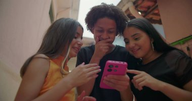 Gençler banliyö sokaklarındaki akıllı telefon ekranına gülüyor ve işaret ediyor, eğlence ve etkileşimli an arkadaşlığı, dijital bağlılığı ve gençlik neşesini gösteriyor