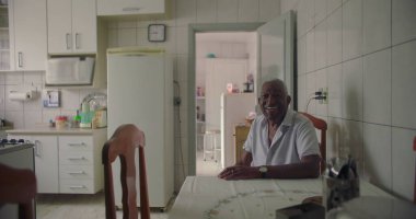 Bir yaşlı siyah adam mutfak masasında oturmuş, sıcak bir şekilde gülümsüyor, pozitiflik ve neşe saçıyor, fayanslarla çevrili duvarlar, buzdolabı ve rahat bir evde ahşap sandalyelerle.