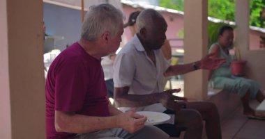 Avluda yemek tabakları, rahat toplanma, sosyal etkileşim, nesiller arası topluluk, tropikal ortam tutarken yaşlı erkekler sohbet ediyorlar.