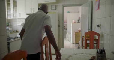 Bir yaşlı siyah adam mutfak masasında oturmuş düşünceli ve düşünceli bir yüz ifadesiyle, döşeli bir iç döşeme, buzdolabı ve bir ev düzeninde ahşap sandalyelerle çevrili.