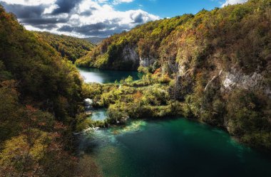 Hırvatistan 'ın Plitvice Lakes Ulusal Parkı' ndaki yemyeşil tepeler ve sonbahar yapraklarıyla çevrili turkuaz göllerin nefes kesici manzarası. Dramatik gökyüzü bu çarpıcı doğal manzaraya derinlik katar.