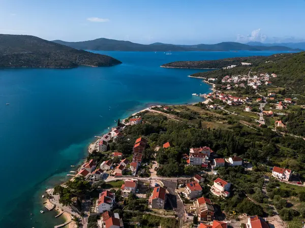 Açık mavi denizi, canlı yeşilliği ve geleneksel kırmızı çatılı evleri olan bir Hırvat sahil kasabasının çarpıcı görüntüsü. Gerçek bir Akdeniz cenneti