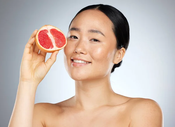 Grapefruit, skincare and woman with food for face, wellness and beauty against a grey mockup studio background. Portrait of young, happy and Asian model with fruit for nutrition, diet and body health.