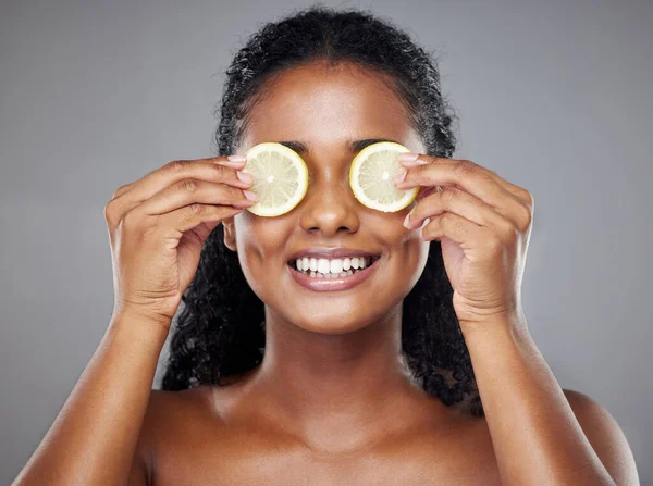 Skincare, portrait and woman with lemon on her eyes for health, beauty and wellness standing in a studio. Happy, smile and face of girl model from Brazil with citrus fruit isolated by gray background.