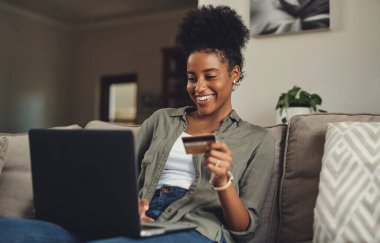 Çevrimiçi alışveriş en iyisidir. Evde bir kanepede dinlenirken kredi kartını ve laptopunu kullanan güzel bir genç kadın.
