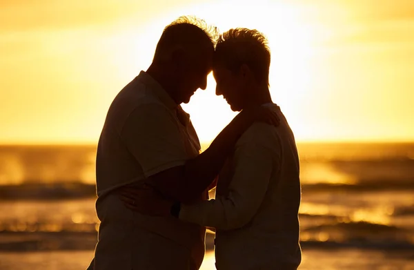 Couple, elderly and silhouette at beach with hug in sunset, evening or dusk by water, waves or horizon together. Senior, man and woman by ocean, sea or sunshine for care, affection or love in romance.