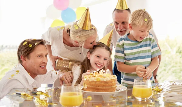 Girl, birthday cake and family celebrating a children event with food and a party outdoor. Happy smile of kids, father and mom kiss to celebrate youth and kid event with happiness, love and confetti.