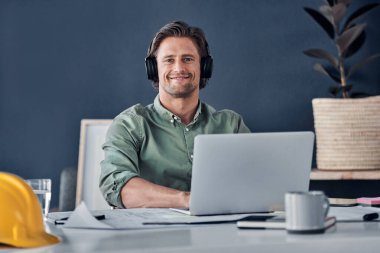 Everything is working in my favor today. Portrait of a handsome young architect wearing headphones while working in his office clipart