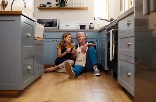 Couple, senior and wine in kitchen, floor and wood for romance, bonding or love in home for happiness. Elderly, man and woman make toast, happy or smile for glass, drinking and together on flooring.