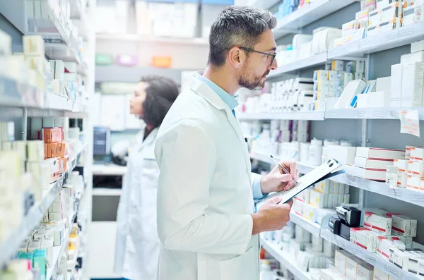 If it makes you better, hell find it. a mature man doing inventory in a pharmacy with his colleague in the background