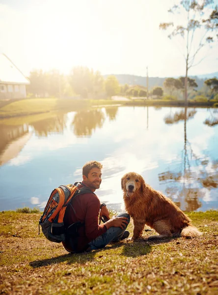 他喜欢呆在外面一个英俊的年轻人和他的狗坐在公园湖边的全长画像 — 图库照片