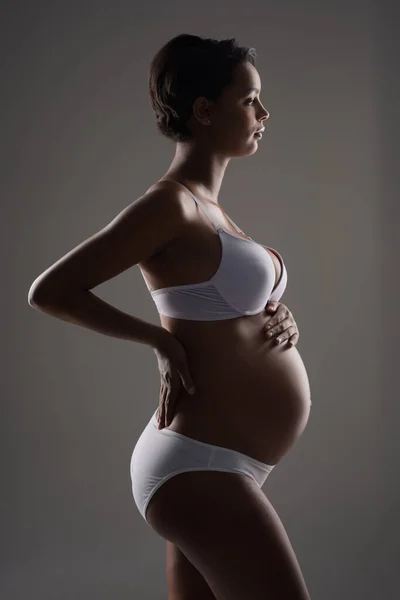 出産前から母親としての生活が始まる スタジオショットのA美しいです若いです妊娠中の女性のポーズで下着に灰色の背景 — ストック写真