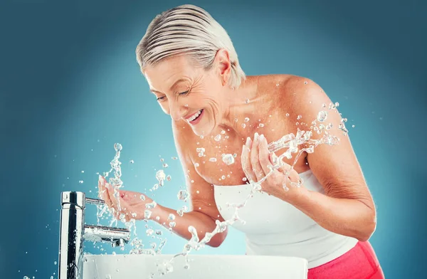 Beauty splash, senior and woman cleaning face in studio for wellness, skincare and hygiene on blue background mockup. Water splash, senior woman and happy, smile and water, beauty and skin facial.