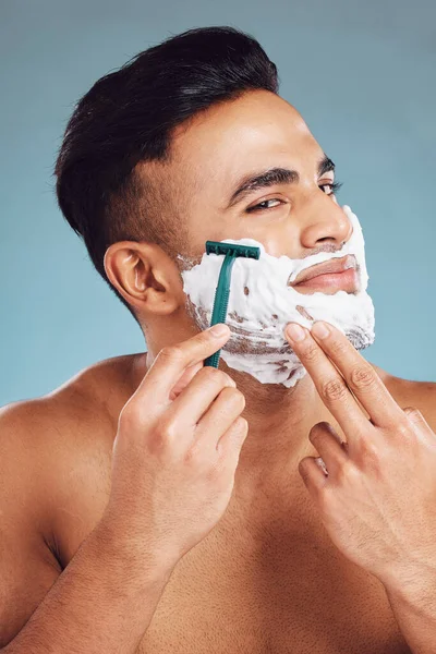 Beauty, skincare and man shaving beard for clean face for dermatology, health and wellness on blue studio background. India male model with foam for healthy skin after facial shave and self care.
