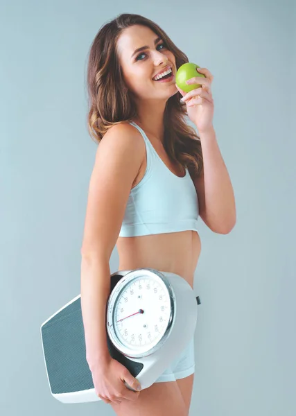 健康的な食事とスケールはあなたを幸せにします 灰色の背景をしたスケールとリンゴを持った健康な若い女性のスタジオショット — ストック写真