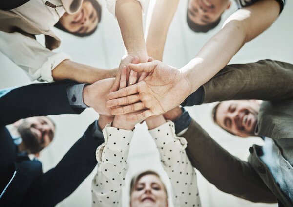 Aiming for success . Low angle shot of a group of cheerful businesspeople forming a huddle with their hands and looking down inside of the office