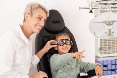 Çocuk, göz muayenesi ve göz doktoruyla göz sağlığı danışmanlığı sırasında gözlük için görüş testi yapıyorlar. Göz tedavisiyle mutlu bir kız. Optometri uzmanı. Hastanın sağlığına ve sağlığına yardım ediyor..