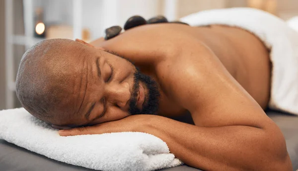 Black man, spa bed hot stone massage and relax body, treatment and luxury physical therapy for health, calm and wellness. Zen man, rest salon and rock for healthcare, recovery or skincare on back.