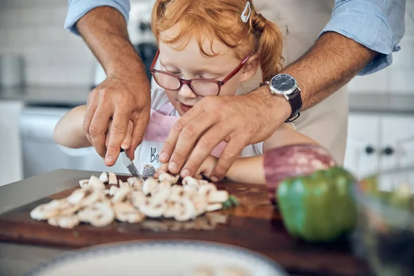 Kid, dad and cooking, cutting board and food in kitchen, family home or house for childhood fun, learning and development. Parent helping little girl chop mushroom vegetables for lunch or dinner meal.