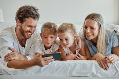 Çocuklu mutlu bir aile, telefon ve evde yatakta uzanmış anne, baba ve kız birlikte video izliyorlar. Sevgi, eğlence ve aile baba, anne ve kızlarıyla yatak odasındaki akıllı telefon görüşmesinde.