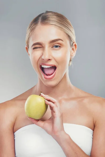 Woman, health and wink portrait with apple for fiber diet, nutrition and wellness lifestyle. Fruit, healthy and digestion dieting marketing with happy model girl on gray studio background