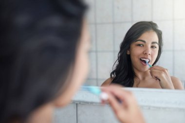 Daha sağlıklı bir gülümsemeye yolculuk, eksiksiz bir temizlik ile başlar. Çekici genç bir kadın banyoda dişlerini fırçalıyor.