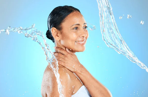 Mature woman, wet or water splash on blue background in studio skincare wellness, hygiene maintenance or body cleaning. Smile, happy or beauty model with pouring liquid healthcare or hydration motion.