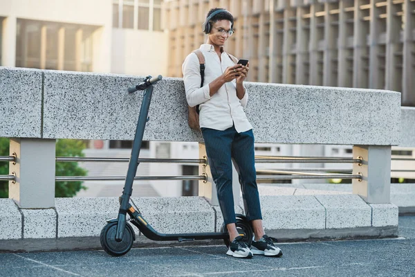 Phone, music and electric scooter with a business black man streaming audio during his commute into work. Mobile, headphones and transport with a male employee typing a text message in an urban town.