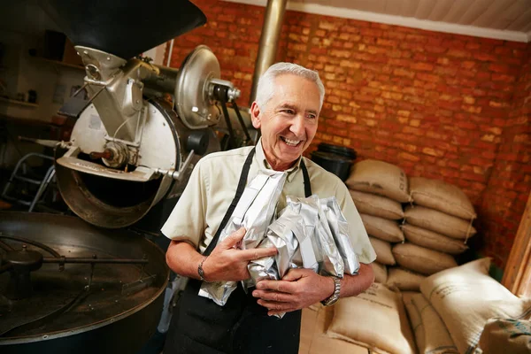 醸造家としての彼の技術を誇りに思う 焙煎所で働きながらコーヒー豆の袋を持った先輩が — ストック写真