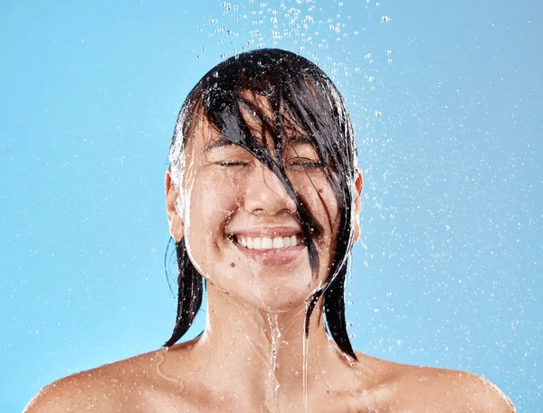 stock image Shower, woman and water with a face cleaning for clean, wash and hygiene on a blue studio background. Bathroom, showering and asian female cleansing her body for health and wellness in studio.