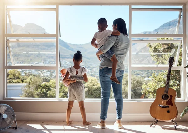 Child, window and view with mother and brother while holding her doll at home. Little girl, woman and siblings with parent looking at while bonding and relaxing with mom in the family home with love.