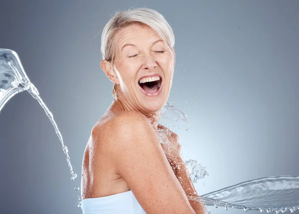 Water splash, health and portrait of senior woman shocked and surprised on gray background. Skincare, wellness and beauty, a happy mature lady splashed with water with happy smile on face in studio